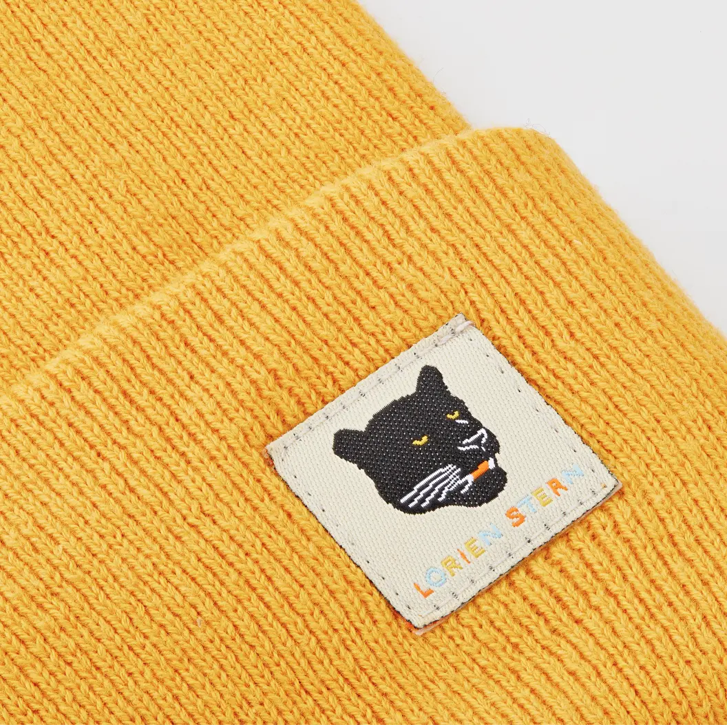 Close-up of a yellow knitted beanie featuring a small black cat patch.