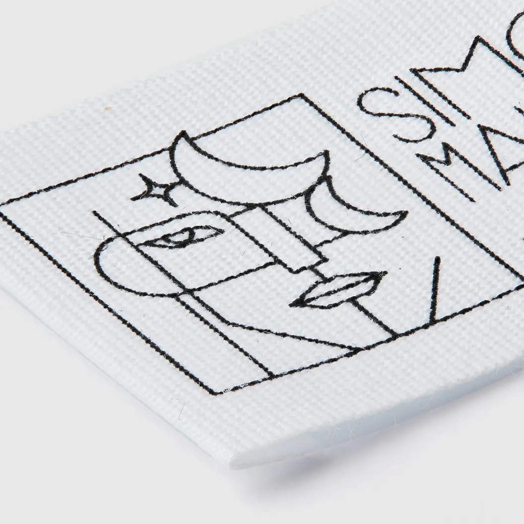 White woven label with a black minimalist line drawing of a face and crescent moon, along with the text “SIMON MATUS.