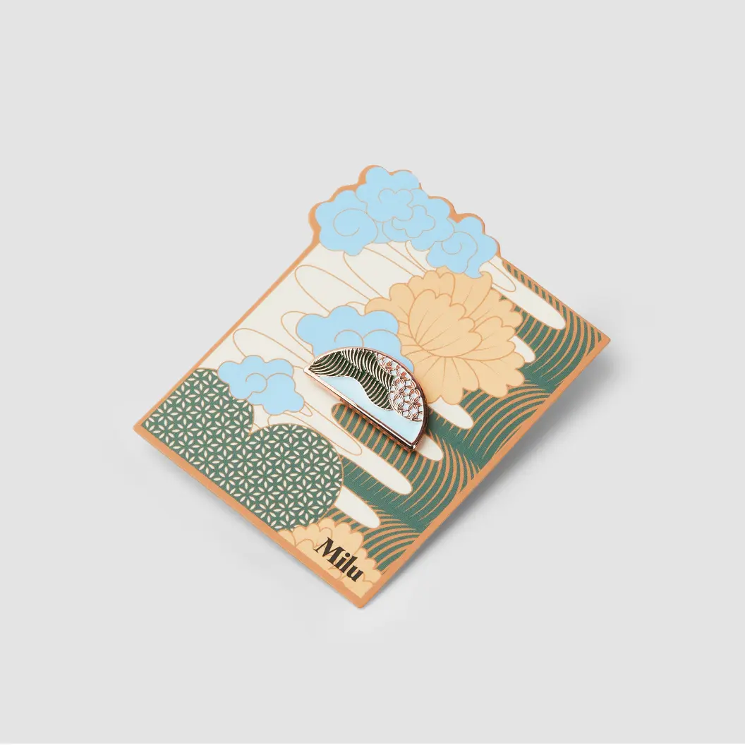 Decorative backer card with floral designs in orange, blue, and green, holding a metal pin in the shape of a leaf.