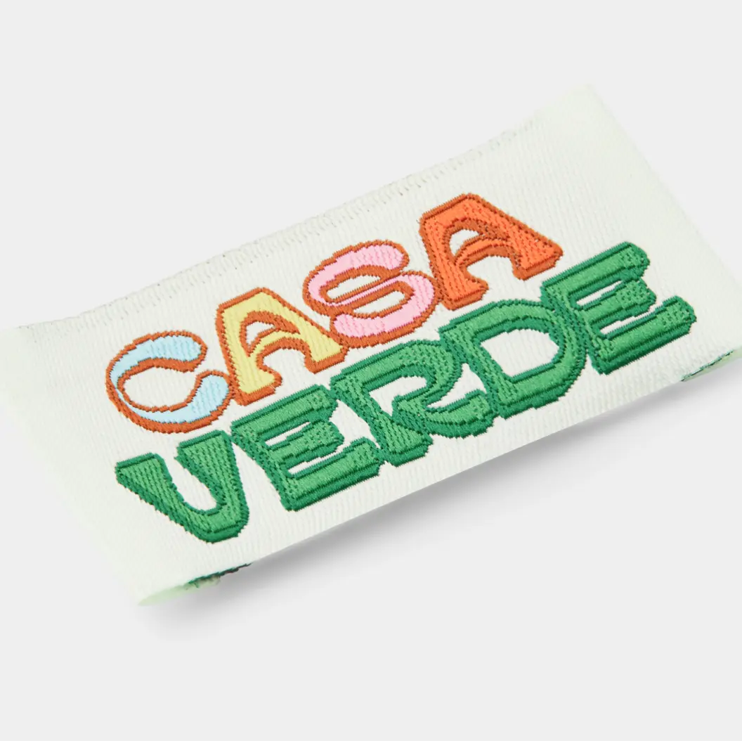 Cream-colored woven label featuring multicolored embroidered text “CASA VERDE” in green, orange, and pink