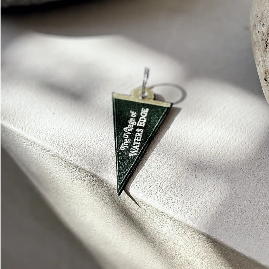 Green pennant embroidered keychain with ‘The Village at Waters Edge’ text, lying on a light surface