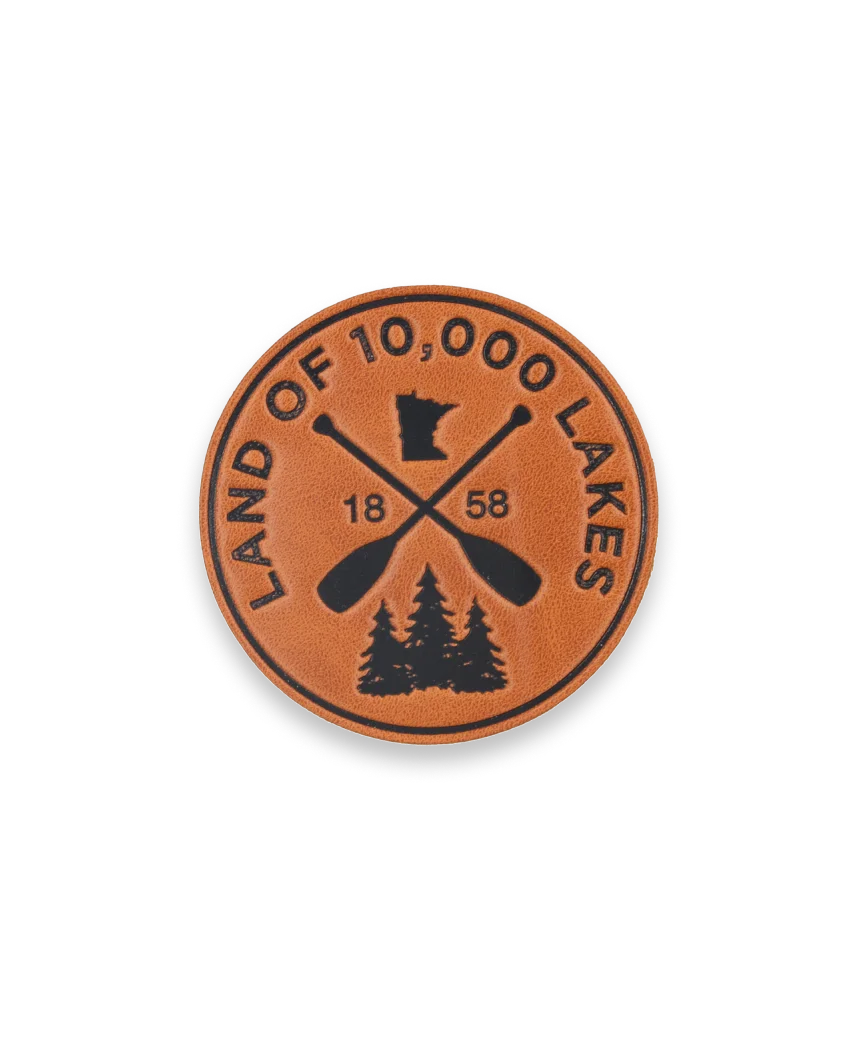 Circular brown leather patch with black design elements, including crossed paddles and “Land of 10,000 Lakes, 1858.”