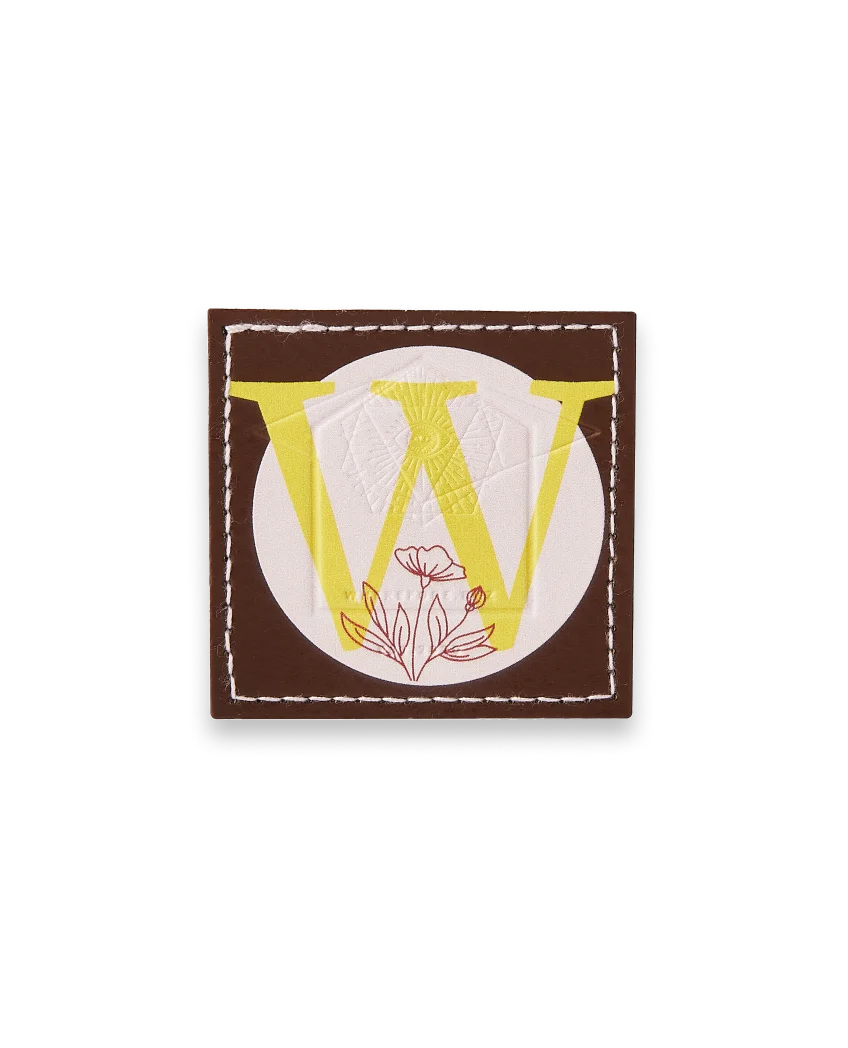 Square leather patch with brown border and bold yellow “W” with floral elements.